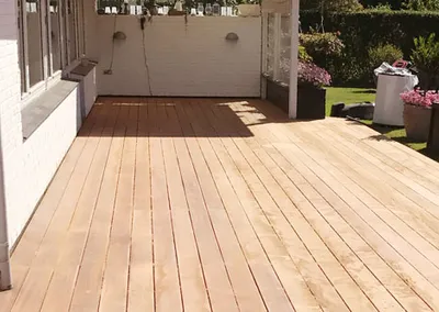Ny terrasse af lyse træbrædder langs et hvidt murstenshus med have ved siden af
