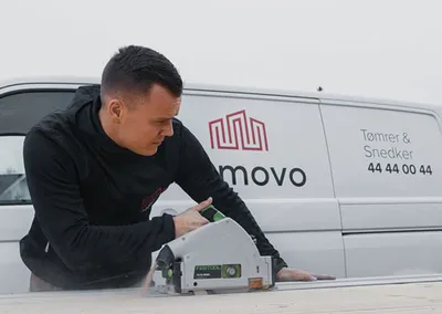 Tømrer fra Nemovo skærer en plade med dyksav foran firmabilen, mærket 'Tømrer & Snedker 44 44 00 44'