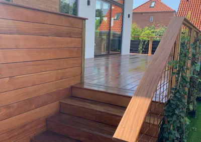 Hævet terrasse i hårdttræ med trappe, gelænder og træbeklædt side ved et hvidt hus