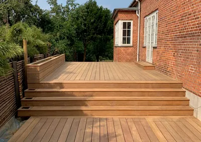 Stor terrasse i lyst hårdttræ i to niveauer med brede trin langs et rødt murstenshus