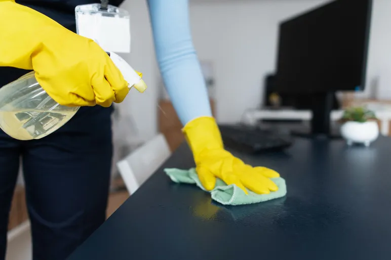 Rengøringsassistent i gule gummihandsker sprayer og tørrer et skrivebord af på et kontor